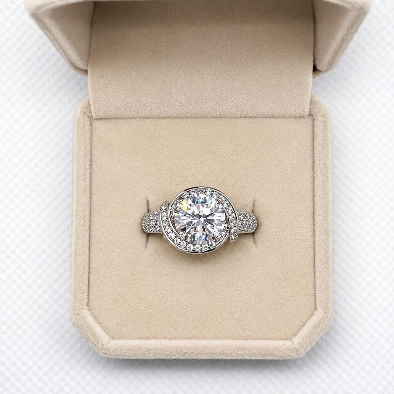 Puede incluir: Un anillo plateado con una piedra preciosa grande, redonda y transparente en el centro, rodeada de piedras m&aacute;s peque&ntilde;as y transparentes. El anillo est&aacute; en una caja beige.