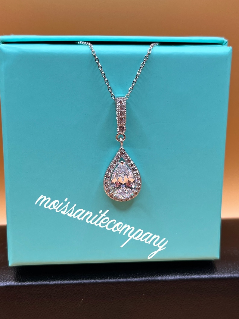May include: A silver necklace with a pear-shaped pendant set with a large clear stone surrounded by smaller stones. The necklace is in a turquoise box with the text "moissanitecompany" printed on the bottom.