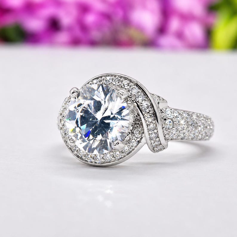 Puede incluir: Un anillo plateado con una piedra preciosa grande, redonda y transparente en el centro, rodeada de piedras m&aacute;s peque&ntilde;as y brillantes. La banda tambi&eacute;n est&aacute; incrustada con piedras m&aacute;s peque&ntilde;as, creando un efecto deslumbrante. El anillo est&aacute; sobre un fondo suave y borroso.