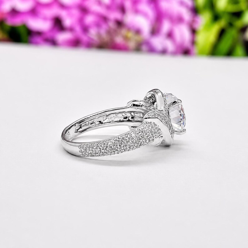 Puede incluir: Un anillo plateado con una piedra preciosa grande y transparente en el centro. La banda est&aacute; incrustada con peque&ntilde;as piedras brillantes. El anillo est&aacute; sobre un fondo blanco con flores rosas borrosas en el fondo.