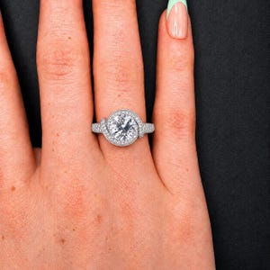 Puede incluir: Primer plano de un anillo de diamantes con una gran piedra central redonda. El anillo est&aacute; engastado en una banda plateada y rodeado de diamantes m&aacute;s peque&ntilde;os. El anillo est&aacute; en una mano, con fondo negro.