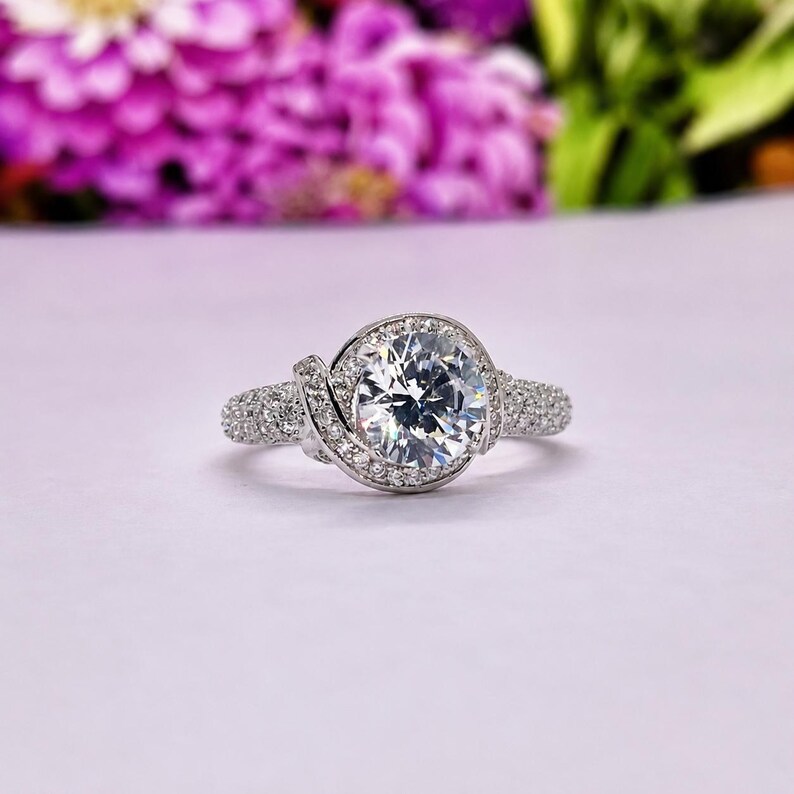 Puede incluir: Un anillo plateado con una piedra preciosa grande, redonda y transparente como pieza central. La piedra preciosa est&aacute; rodeada de piedras m&aacute;s peque&ntilde;as y transparentes. La banda tambi&eacute;n est&aacute; incrustada con piedras m&aacute;s peque&ntilde;as. El anillo est&aacute; sobre un fondo rosa suave con flores moradas borrosas.
