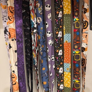Op de afbeelding: Een verzameling Halloween-thema lanyards met verschillende patronen, waaronder pompoenen, vleermuizen, geesten en Jack Skellington. De lanyards zijn gemaakt van stof en hebben metalen clips en ringen. Het kleurenpalet omvat oranje, paars, zwart en blauw.