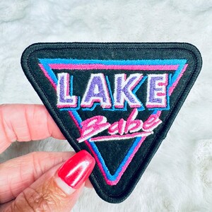 May include: A black triangular patch with pink and blue neon-style trim. The patch says "LAKE Babe" in purple and pink lettering.