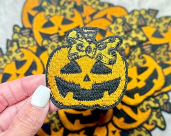 May include: A close-up of a yellow and black embroidered pumpkin patch iron-on patch with a black bow and a black and yellow butterfly design. The patch is being held by a hand.