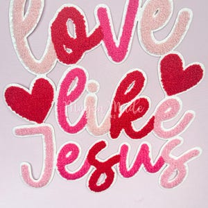 Puede incluir: Parche bordado "Love like Jesus" en rosa, rojo y blanco con una textura peluda. El parche presenta dos corazones.