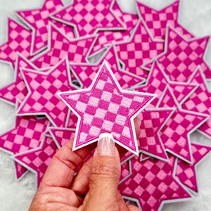 Peut inclure: Un tas de patchs en forme d'étoile à carreaux rose et blancs. Les patchs sont en tissu et ont un contour blanc. Les patchs sont disposés en pile, le patch du dessus étant tenu par une main.