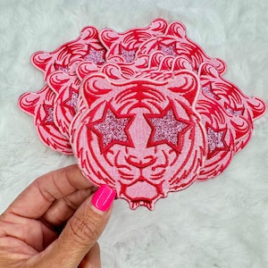 May include: A stack of pink iron-on patches featuring a tiger face with pink glitter star eyes.