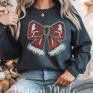 Op de afbeelding: Een donkergrijs sweatshirt met een rode en witte pailletten strik met een gouden gesp. De strik heeft een witte bontkraag en is versierd met kleine witte bellen. Het sweatshirt heeft de tekst "Magen Made Designs" onderaan.