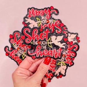 Puede incluir: Un conjunto de parches termoadhesivos negros y rojos con purpurina y un diseño de cupido rosa y blanco. Los parches tienen el texto "Shot through the heart".
