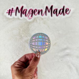 May include: A silver iridescent disco ball patch with a grid pattern. The patch has a white backing and is held in a hand. The text #MagenMade is visible in the background.