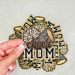 May include: Gold glitter iron-on patch with a black and white pom pom, a gold glitter megaphone, and the text "cheer mom".