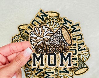 May include: Gold glitter iron-on patch with a black and white pom pom, a gold glitter megaphone, and the text "cheer mom".