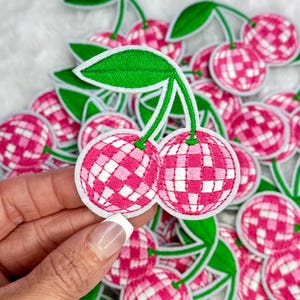 May include: A close-up of a pink and white checkered cherry with a green stem. The cherry is a patch and is surrounded by other similar patches.