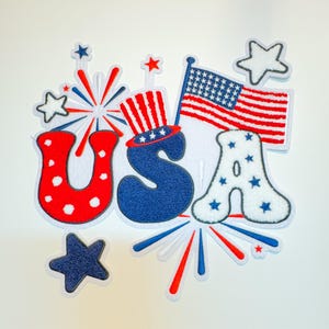 May include: A red, white, and blue felt applique of the word "USA" with stars and fireworks. The "U" is red with white polka dots, the "S" is blue with white stars, and the "A" is white with blue stars. There is a small American flag above the word.