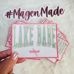 May include: A stack of white iron-on patches with pink and green embroidery. The top patch features the text "LAKE BABE" in green with a pink canoe graphic. The text "#MagenMade" is visible in pink on a white background.