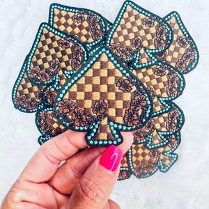 May include: A collection of spade-shaped embroidered patches featuring a brown and tan checkered pattern. Each patch is accented with dark orange floral embroidery and outlined with a bright turquoise stitched border on a black fabric base.