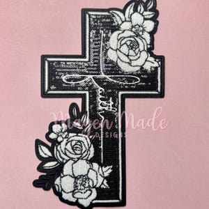 Pode incluir: Etiqueta de ferro bordada em preto e branco com uma cruz com detalhes florais. A cruz é coberta de lantejoulas e as flores são brancas.