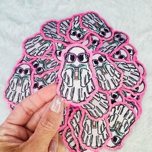 May include: A stack of pink and white felt ghost patches with black outlines. Each ghost is wearing glasses and holding a book.