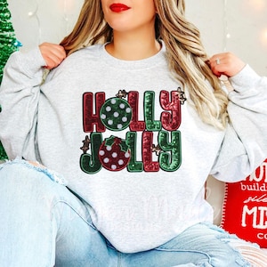 Può includere: Una felpa grigia con un design di paillettes rosse, verdi e bianche che dice "Holly Jolly".