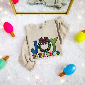 以下が含まれることがあります： JOY TO THE WORLDと書かれたグラフィックプリントが特徴のベージュのクルーネックトレーナー。文字のOは、カラフルなオーナメントで飾られ、大きな赤いリボンがトップにあしらわれた緑色のクリスマスリースに置き換えられています。テキストはマルチカラーのフォントです。