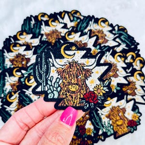 May include: A black felt patch with an embroidered image of a Highland cow with a crescent moon and stars above it. The cow is surrounded by cacti, flowers, and leaves.