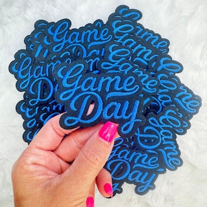 May include: A stack of black and blue iron-on patches with the text "Game Day" in a stylized font.