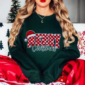 Könnte beinhalten: Grüner Weihnachtspullover mit einem rot-weiß karierten "Frohe Weihnachten"-Motiv. Das Motiv enthält eine Weihnachtsmütze.