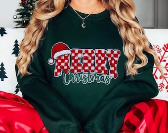 Peut inclure: Sweat-shirt de Noël vert avec un motif "Joyeux Noël" à carreaux rouges et blancs. Le motif comprend un bonnet de Père Noël.