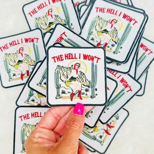May include: A stack of white iron-on patches with a black border. The patches feature a cartoon image of a woman in red boots sitting on a cactus with the text "THE HELL I WON'T" in red and black lettering.