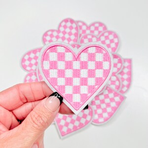 Peut inclure: Un ensemble de patchs en forme de cœur à carreaux rose et blancs. Les patchs sont en feutre et ont un contour blanc.