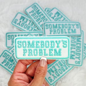 Könnte beinhalten: Ein Stapel türkiser und weißer Bügelbilder mit dem Text "Somebody's Problem" in Weiß. Die Patches sind in einem Stapel angeordnet, wobei ein Patch in einer Hand gehalten wird.