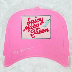 May include: A bright pink baseball cap featuring a rectangular patch with the embroidered red text Spicy Marg Queen and a small light blue chilli pepper icon. The text Magen Made Designs is printed in white on the lower left side of the cap.