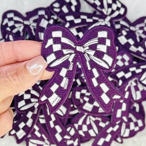 May include: A purple and white embroidered bow patch featuring a checkered pattern. The bow has a central knot with two loops and two long, flowing tails extending downward. The edges of the patch are finished with a dark purple stitched border.