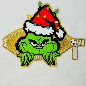 Peut inclure: Personnage vert du Grinch portant un chapeau de Père Noël rouge avec une bordure blanche et des lumières colorées. Le Grinch regarde à travers une pochette zippée en paillettes dorées.