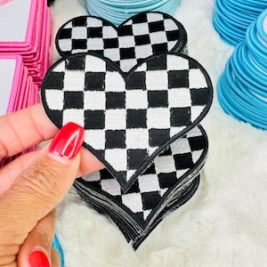 Peut inclure: Un écusson brodé en forme de cœur avec un motif à damier noir et blanc. L'écusson présente une bordure épaisse cousue en noir et fait partie d'une pile d'écussons identiques. Le motif se compose d'une grille de petits carrés.