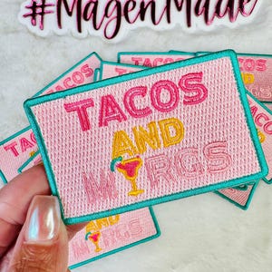 May include: A rectangular embroidered patch with a light pink background and a teal border. The text reads TACOS AND MRGS in pink and yellow thread, with a small yellow margarita glass icon replacing the letter I in the word MRGS.