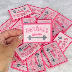 May include: A pile of pink iron-on patches with the text "BARBELL Baddie" and a small barbell graphic.