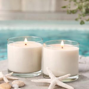 May include: Two white wax candles in clear glass cylindrical containers with lit wicks, accompanied by two white starfish and small smooth pebbles arranged on a flat surface, creating a coastal-themed display with a soft, warm glow.