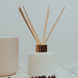 May include: A white ceramic diffuser bottle with a gold lid and wooden diffuser sticks. The bottle is sitting on a marble surface with a sprig of lavender.