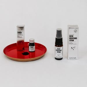 Può includere: Un vassoio circolare rosso contenente un piccolo barattolo nero di polvere Sormeh e la sua scatola argentata, accanto a un flacone di siero nutriente per sopracciglia e la sua scatola argentata. Il flacone ha un erogatore nero e un'etichetta bianca.