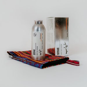 Könnte beinhalten: Eine silberne Flasche mit Körperpeeling-Pulver der Marke "SEFIDAB" mit dem Text "ORIGINAL PERSIAN SKIN DETOX" und "BODY PEEL POWDER" in Englisch und Arabisch. Die Flasche steht auf einem bunten, gestreiften Stoffbeutel.