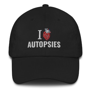 Op de afbeelding: Zwarte baseballpet met witte borduurwerk die "Ik hou van autopsies" zegt met een rood hart en een gestileerde anatomische hartgrafiek.