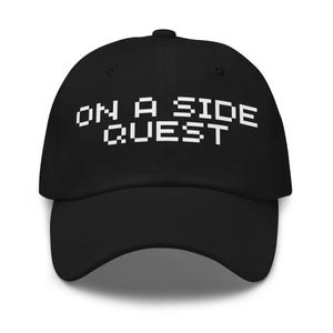 May include: Black baseball cap with the text "ON A SIDE QUEST" in white pixelated font. The cap has a curved brim and a rounded crown. The hat is a classic, casual accessory.