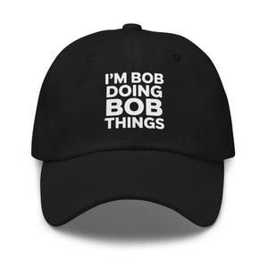May include: Black baseball cap with the text "I'M BOB DOING BOB THINGS" in white block letters. The cap has a curved brim and a button on top. A casual accessory for everyday wear.