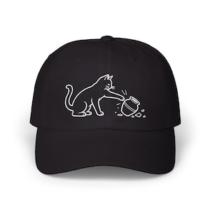 Puede incluir: Gorra de béisbol negra con una ilustración en blanco de un gato y una olla rota. El gato está representado golpeando la olla. La gorra tiene una visera curva y un cierre ajustable.