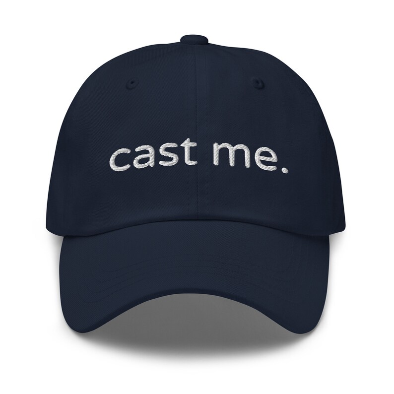 May include: A navy blue baseball cap with white embroidered text that reads "cast me."