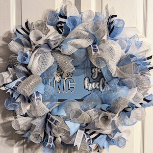 May include: A blue and white wreath with a sign that says "Go Heels" and a UNC logo. The wreath is made of mesh and ribbon.