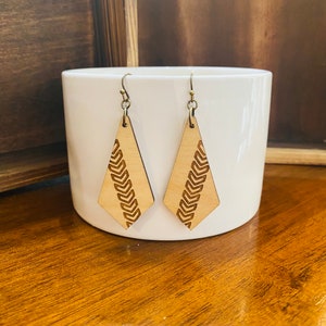 May include: A pair of wooden earrings with a geometric diamond shape and a carved pattern of repeating arrows. The earrings are a light brown colour and have gold-toned hooks.