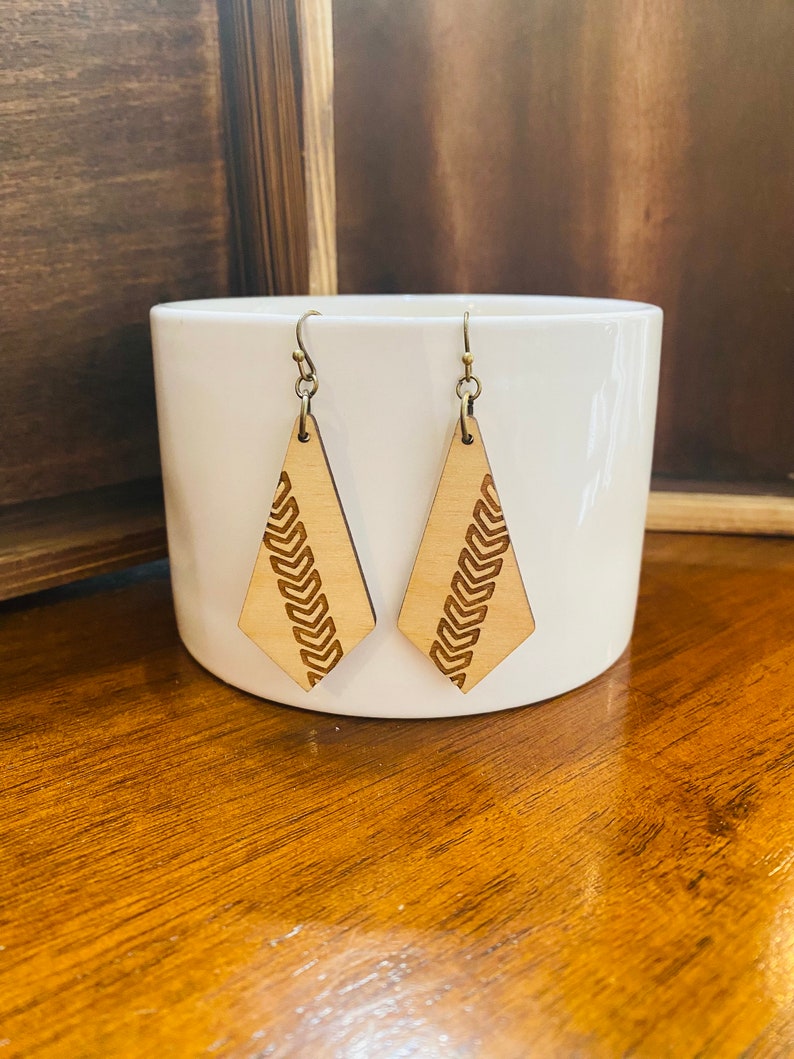 May include: A pair of wooden earrings with a geometric diamond shape and a carved pattern of repeating arrows. The earrings are a light brown colour and have gold-toned hooks.
